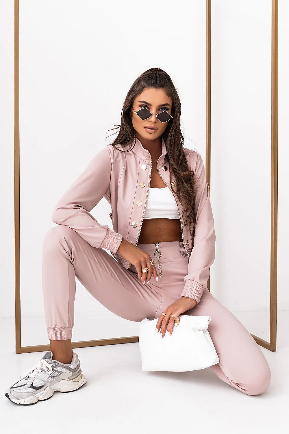 Woman in pink tracksuit with white sneakers and sunglasses, sitting against a white background