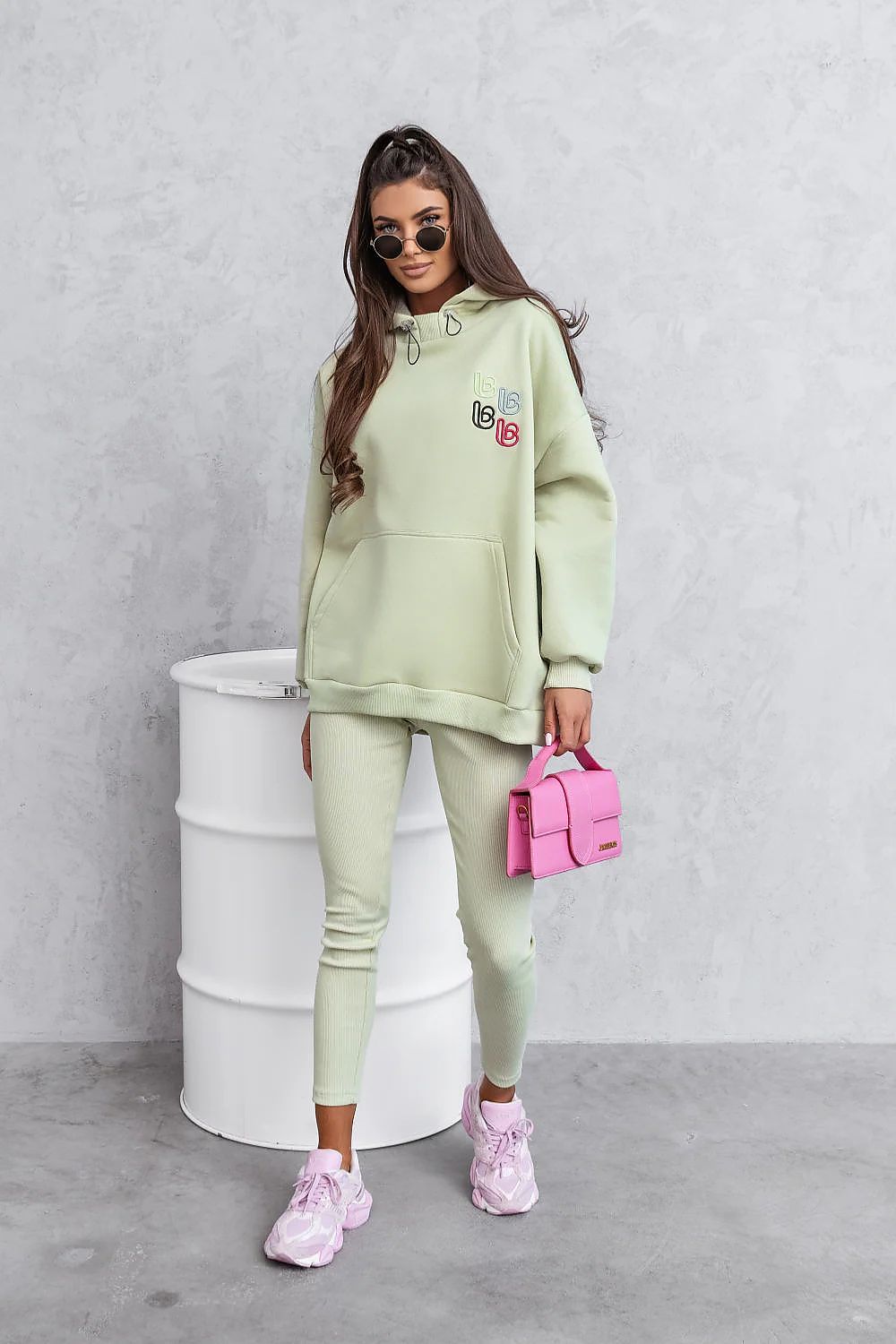 Woman in light green tracksuit with pink accessories standing against a gray wall.