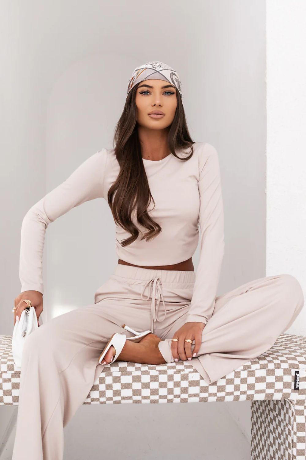 Woman in a beige outfit sitting on a checkered bench against a plain background