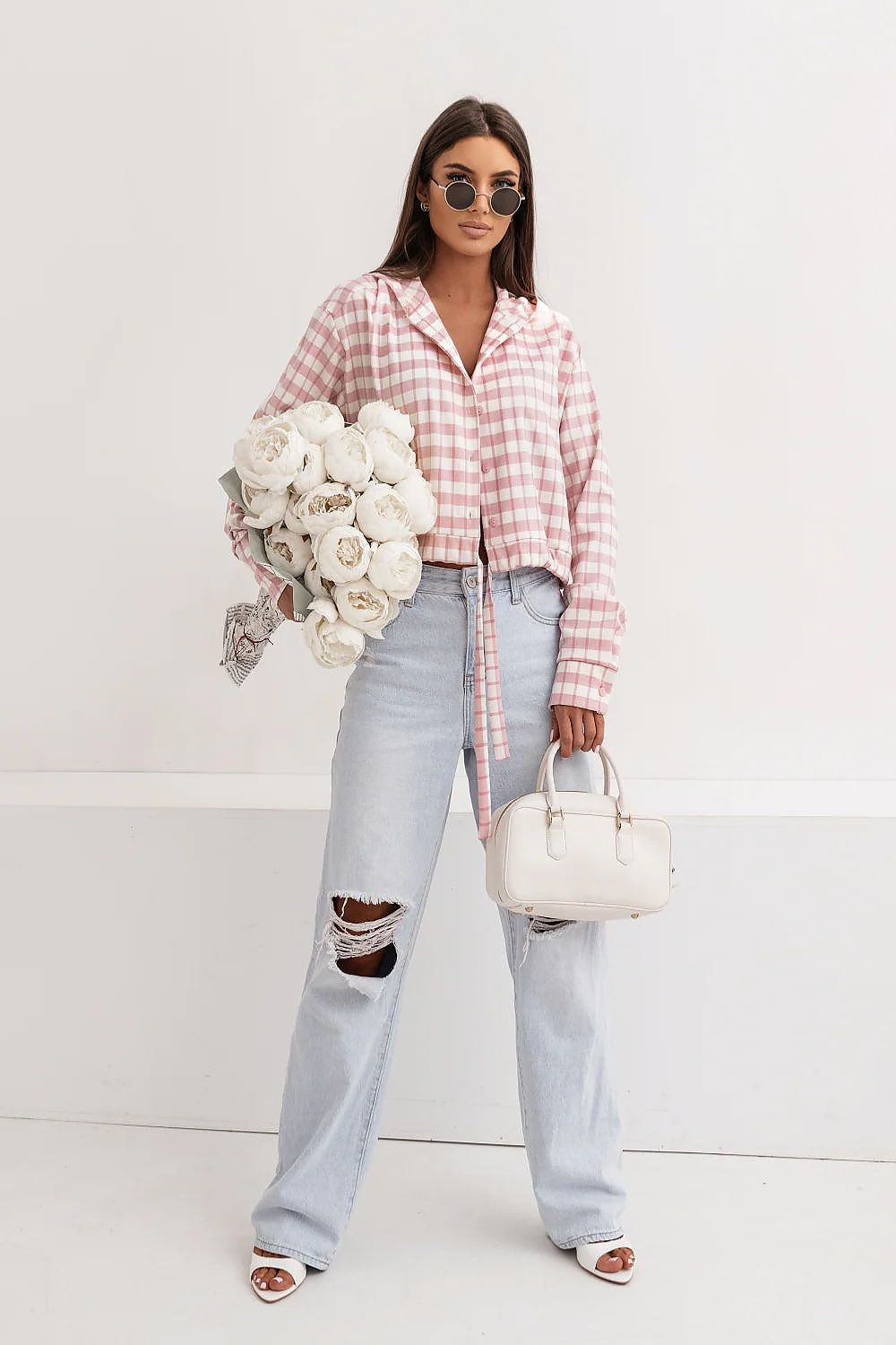Woman wearing a pink plaid shirt and light blue jeans, holding white flowers against a white background