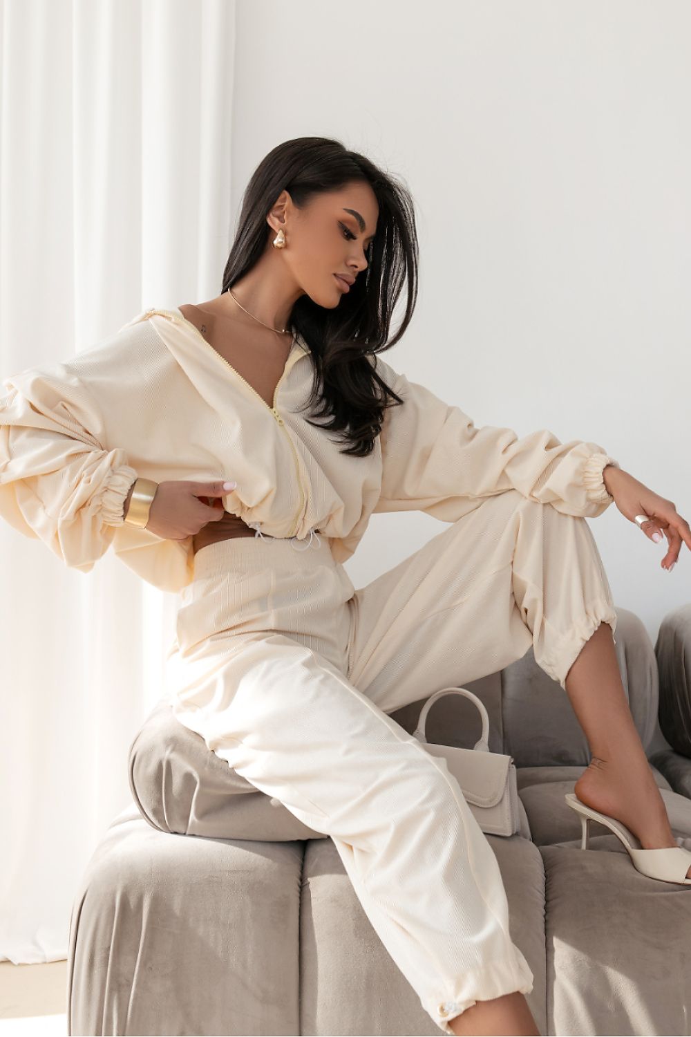 Woman in a cream-colored outfit sitting on a couch in a bright room.