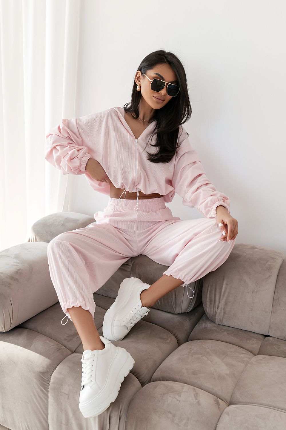 Woman in a pink outfit sitting on a beige sofa with white curtains in the background