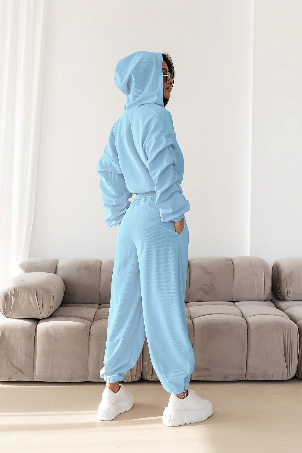 Person wearing a light blue tracksuit standing in a room with a beige sofa.