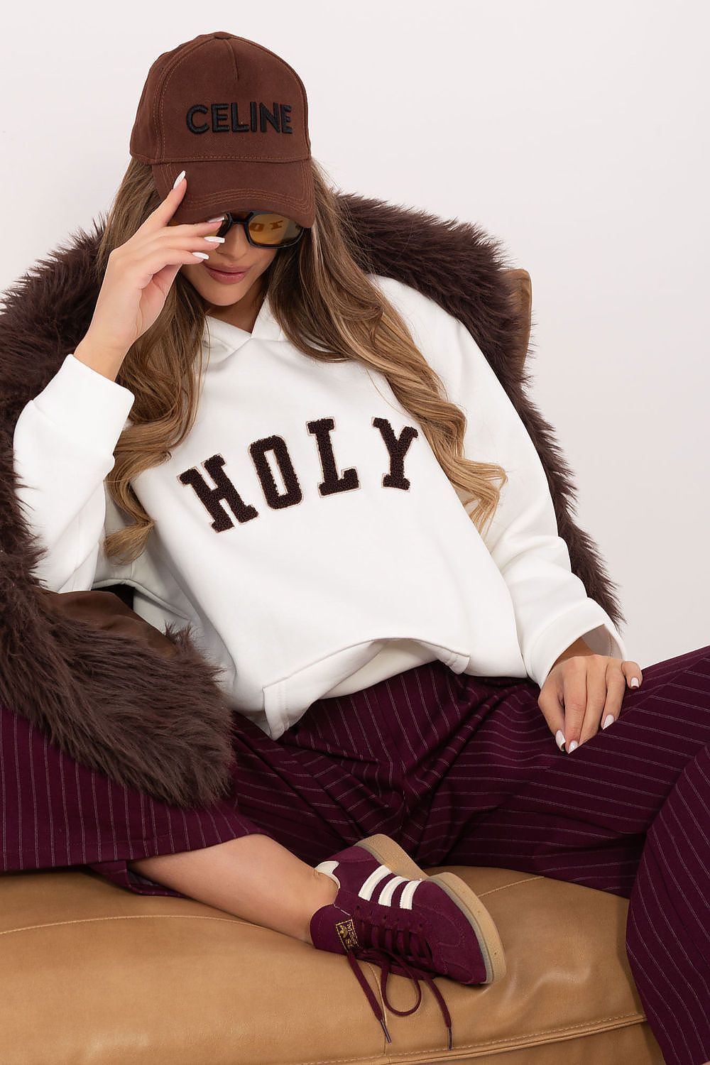 Person wearing a white sweatshirt with 'HOLY' text, maroon pants, and a brown Celine cap, sitting on a brown couch.