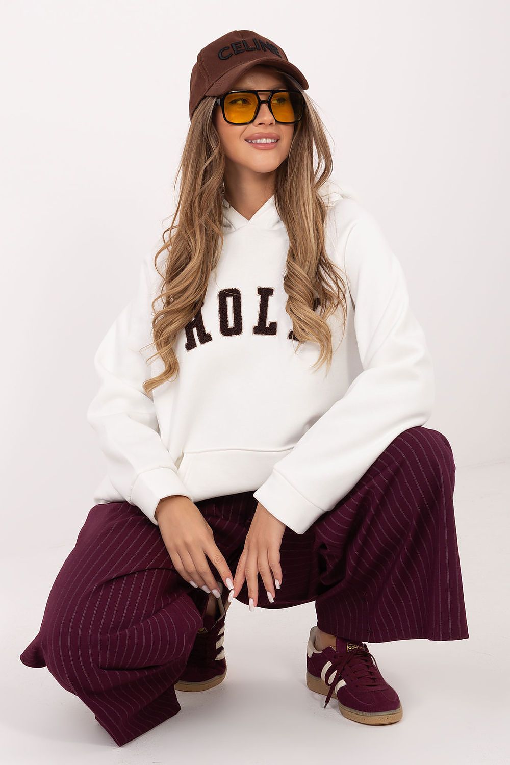 Woman wearing a white hoodie with text, maroon pants, and a cap on a white background