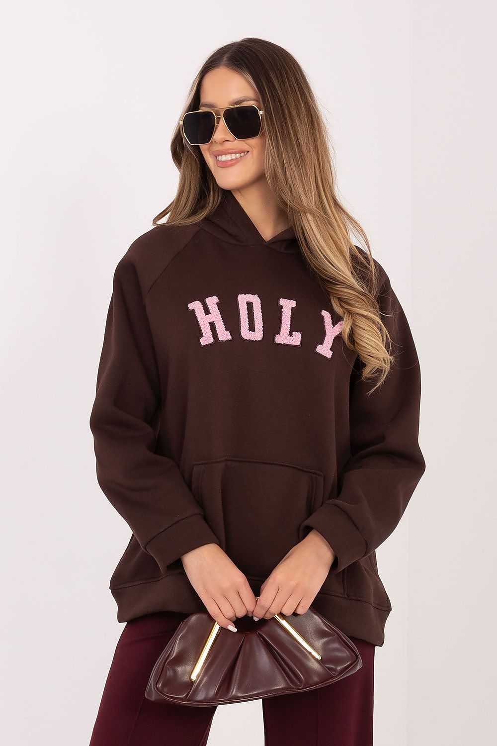Woman wearing a brown hoodie with 'HOLY' text, holding a matching handbag on a white background