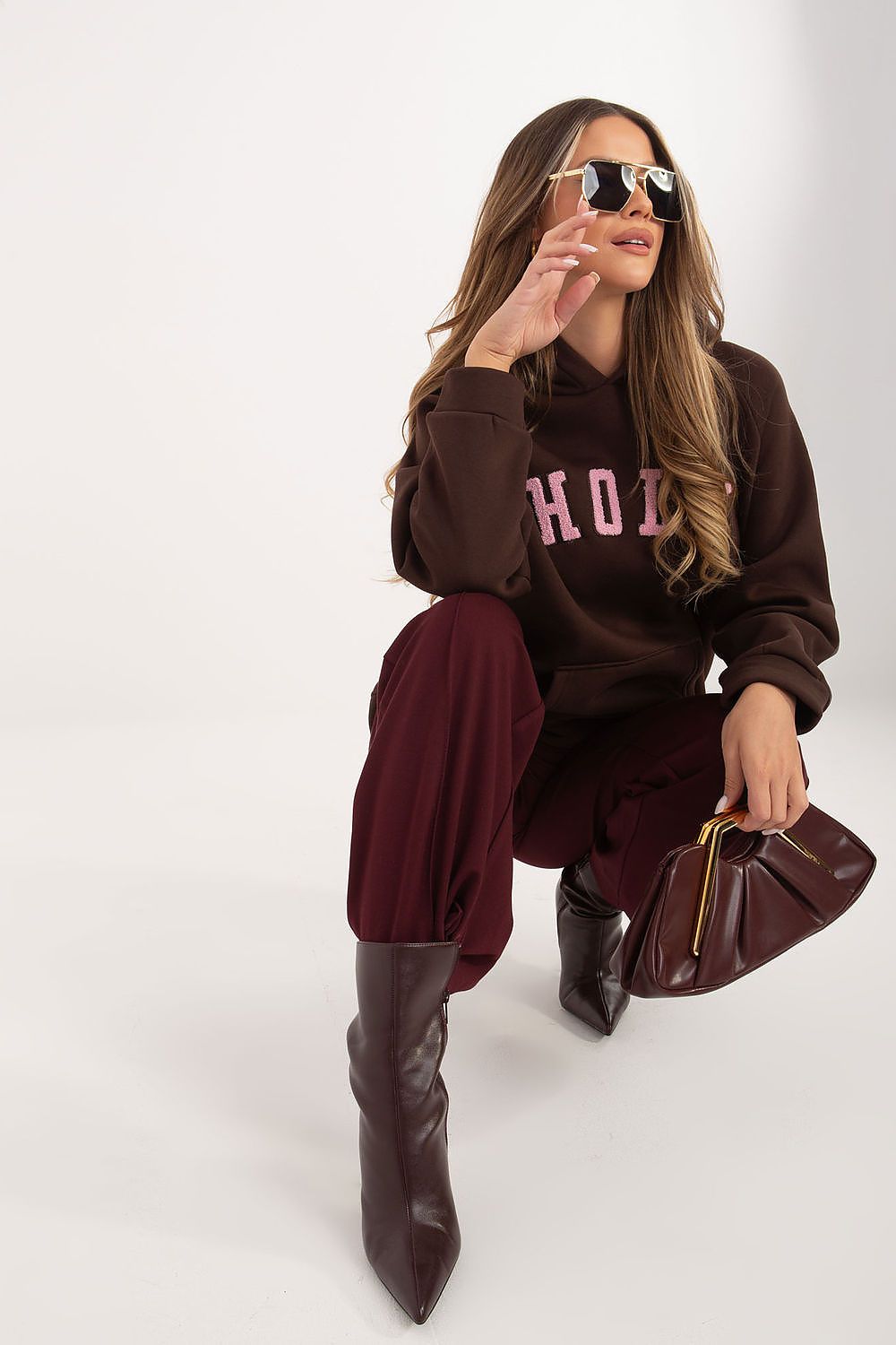Woman wearing a brown hoodie with pink text, burgundy pants, and high-heeled boots, holding a matching handbag on a white background.
