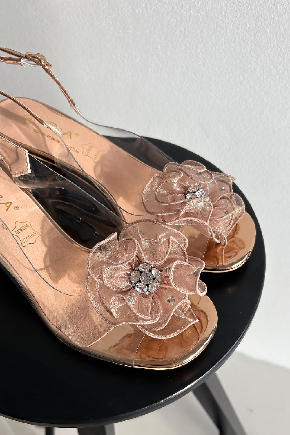 Rose gold sandals with floral designs and clear straps on a black stool against a white background
