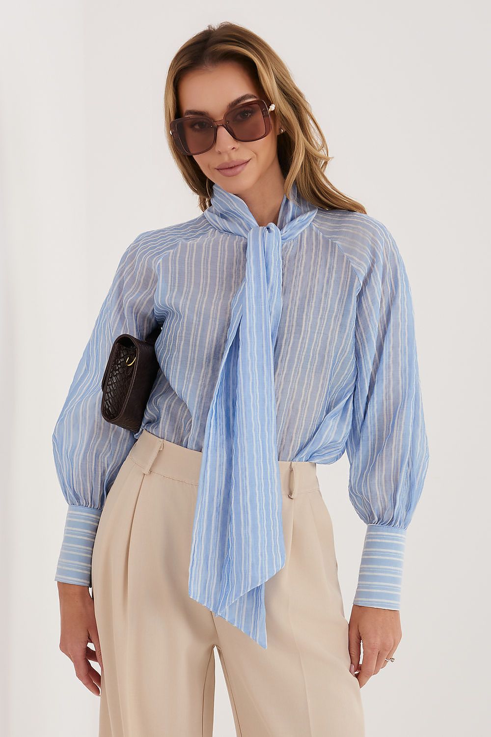 Woman wearing a blue striped shirt with a scarf, sunglasses, and beige pants on a white background