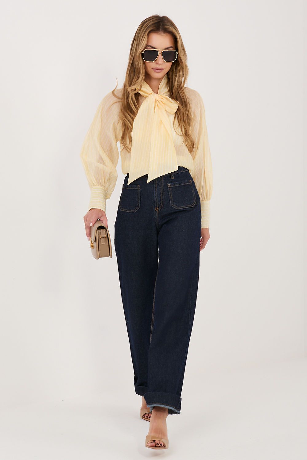 Woman wearing a light yellow blouse, dark blue jeans, and sunglasses on a white background