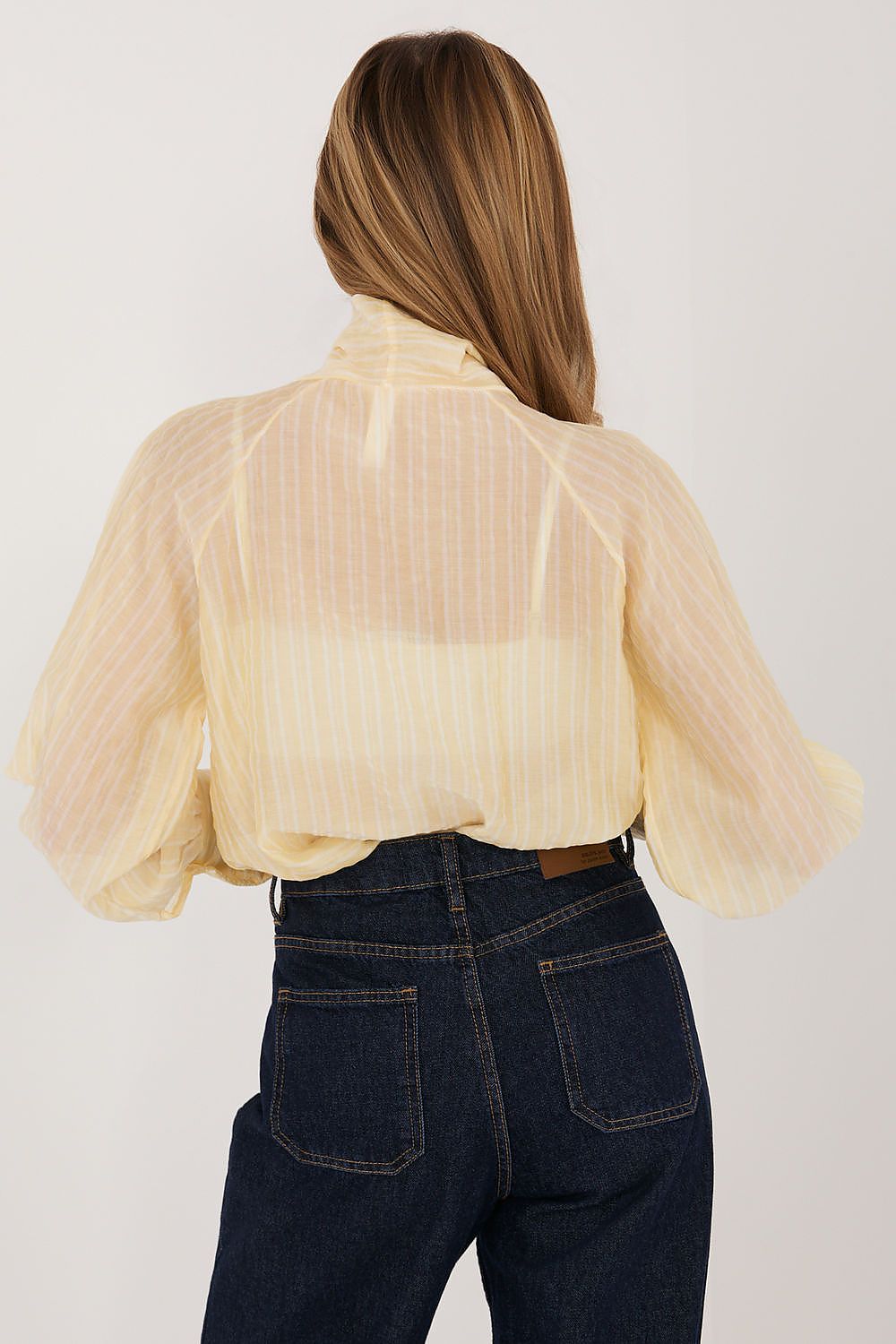 Person wearing a sheer yellow blouse over a white shirt with dark jeans on a light gray background