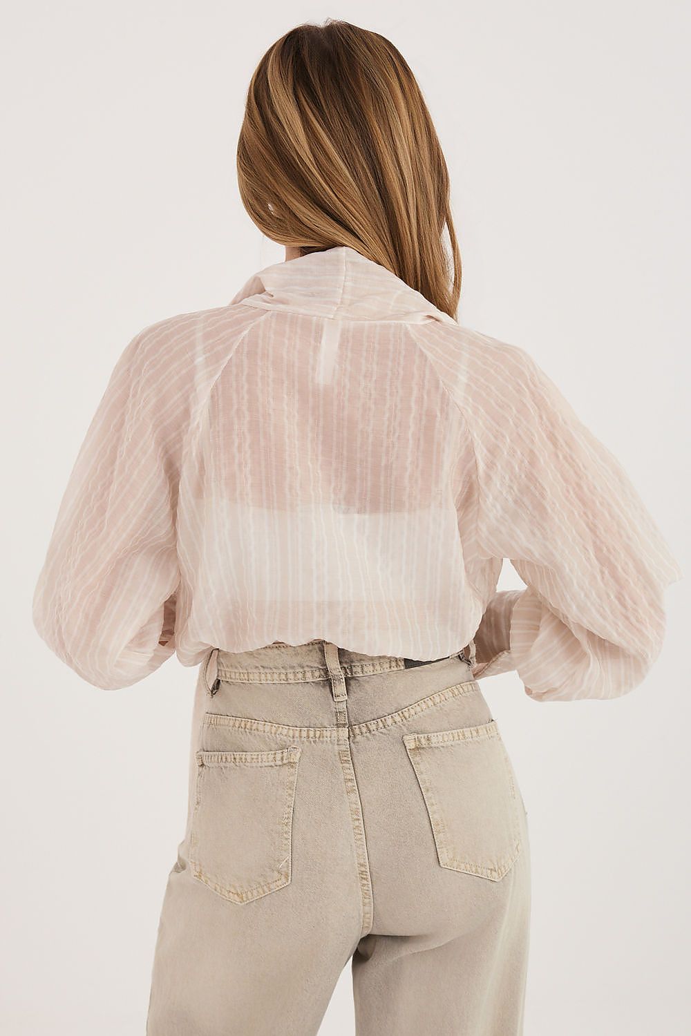 Person wearing a sheer beige blouse over a white shirt with beige pants on a white background