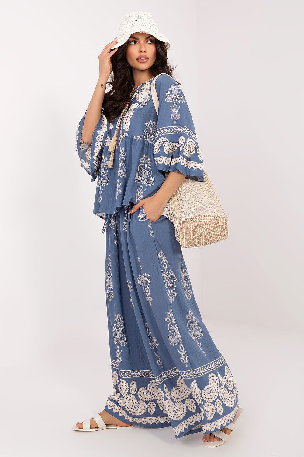 Woman wearing a blue patterned outfit with a white hat and beige bag on a white background