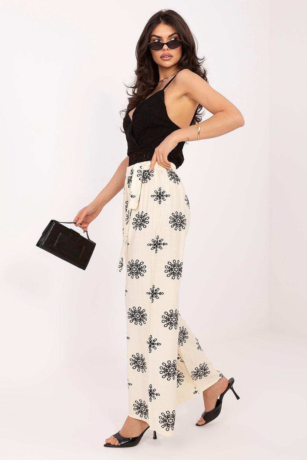 Woman wearing a black top and white pants with black patterns, holding a black clutch against a white background.