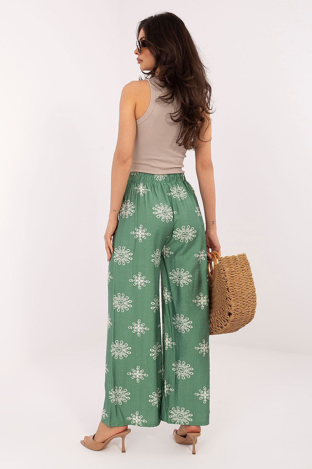 Woman wearing green floral pants with a beige top and holding a straw bag on a white background
