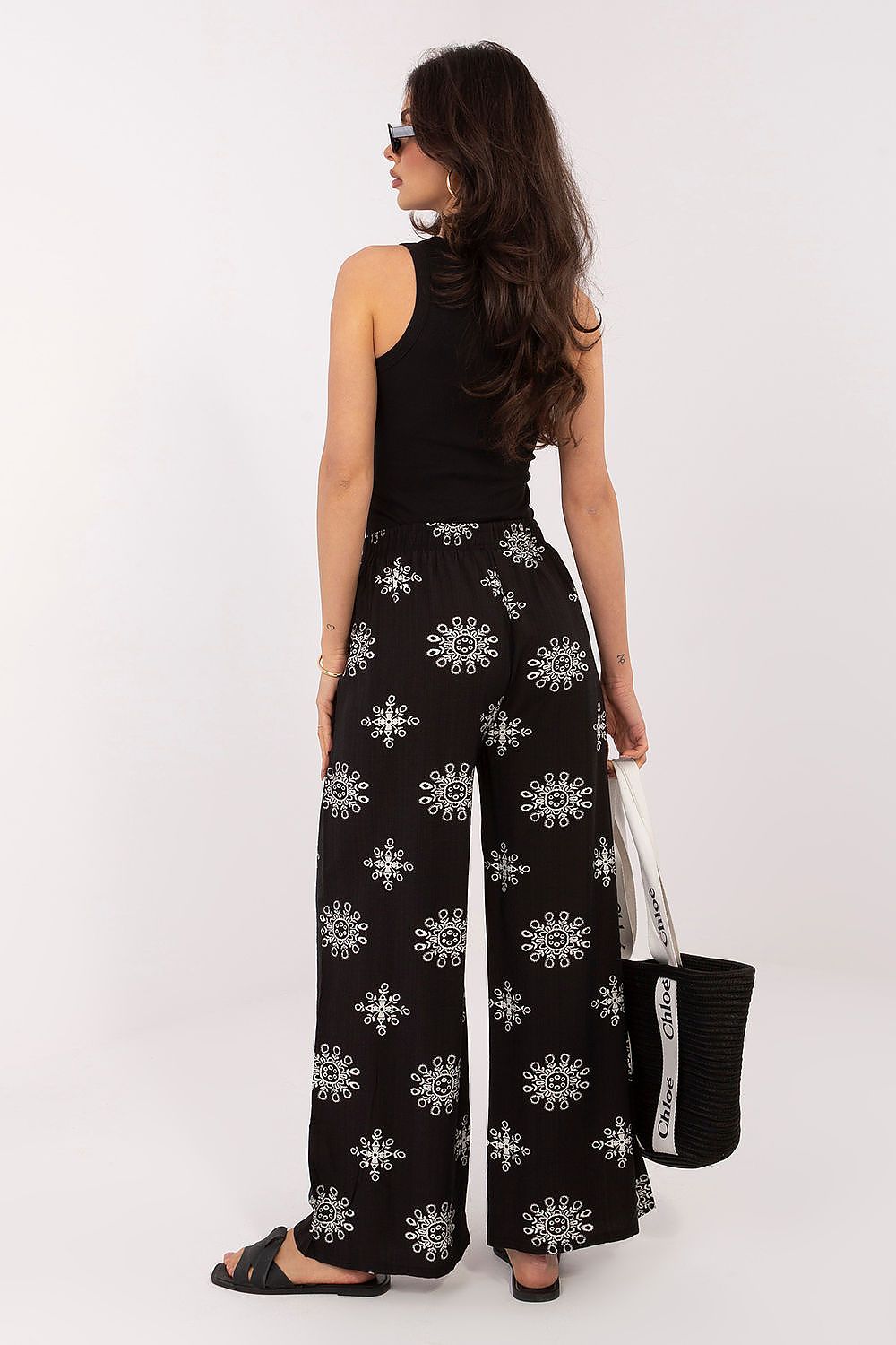 Woman wearing black floral-patterned pants holding a matching bag on a white background