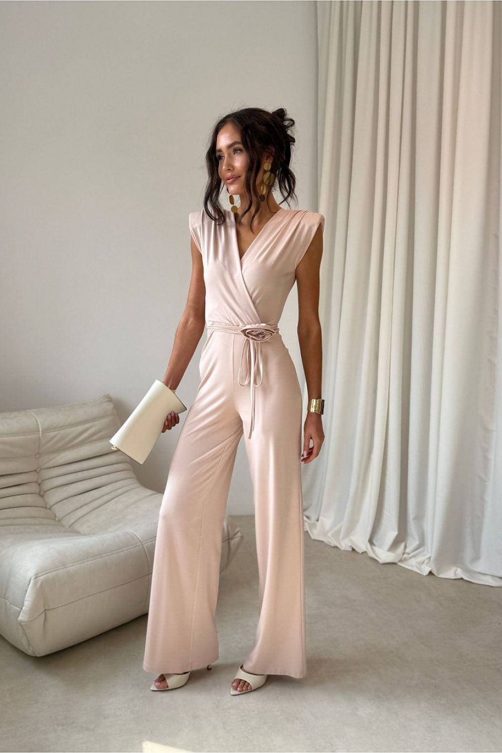 Woman in a pink jumpsuit standing in a room with white curtains and a white couch.