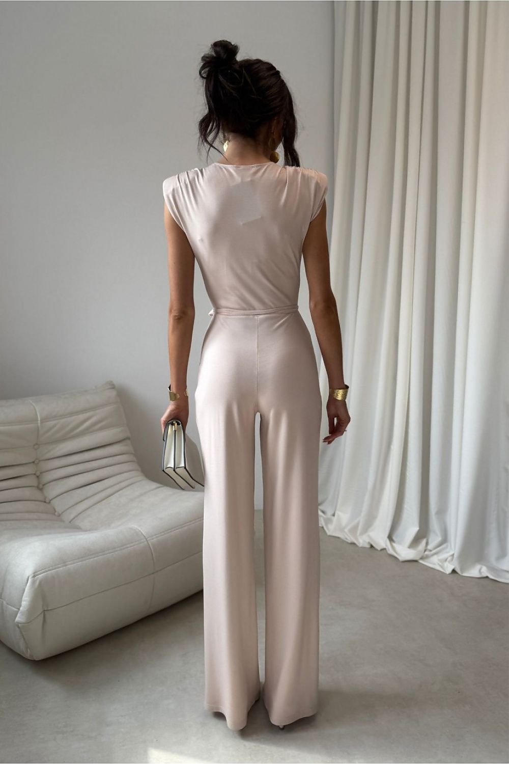 Woman in a beige jumpsuit standing in a minimalistic room with white furniture and curtains.