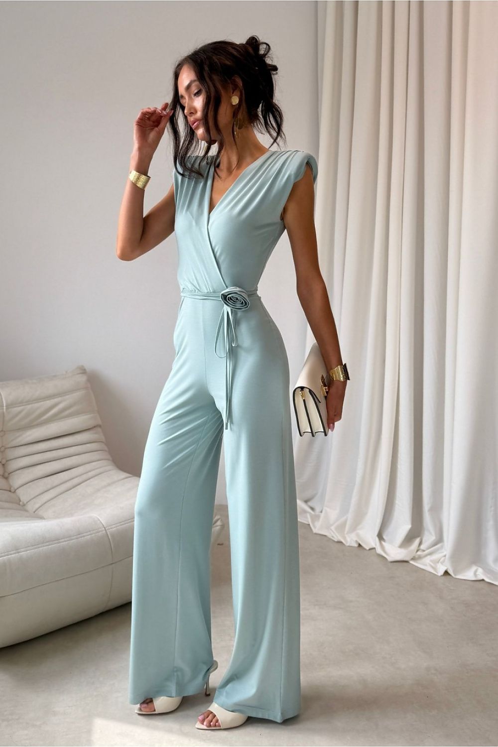 Woman wearing a light blue jumpsuit in a room with white curtains and a white couch.