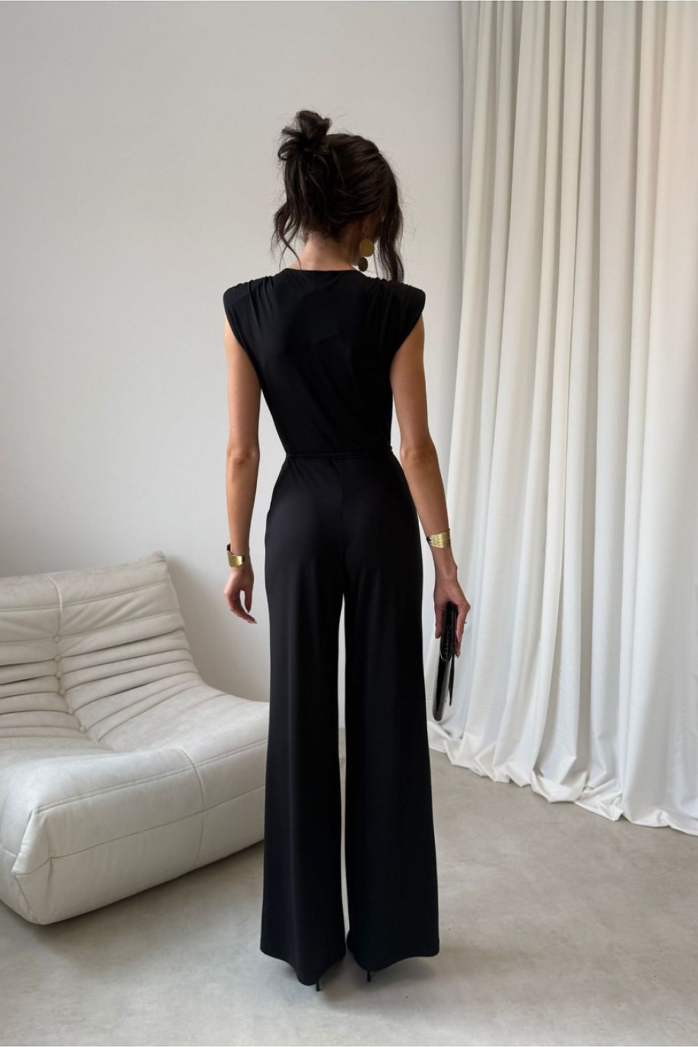 Person wearing a black jumpsuit standing in a room with white curtains and a white couch.