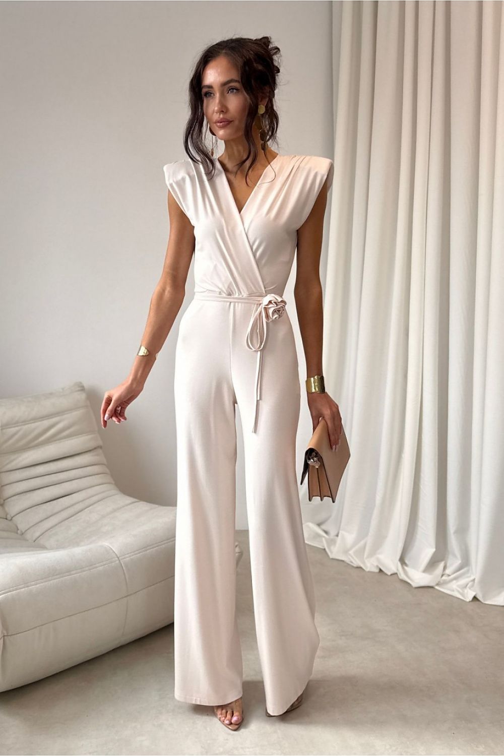 Woman wearing a white jumpsuit in a minimalistic room with a white sofa and curtains.