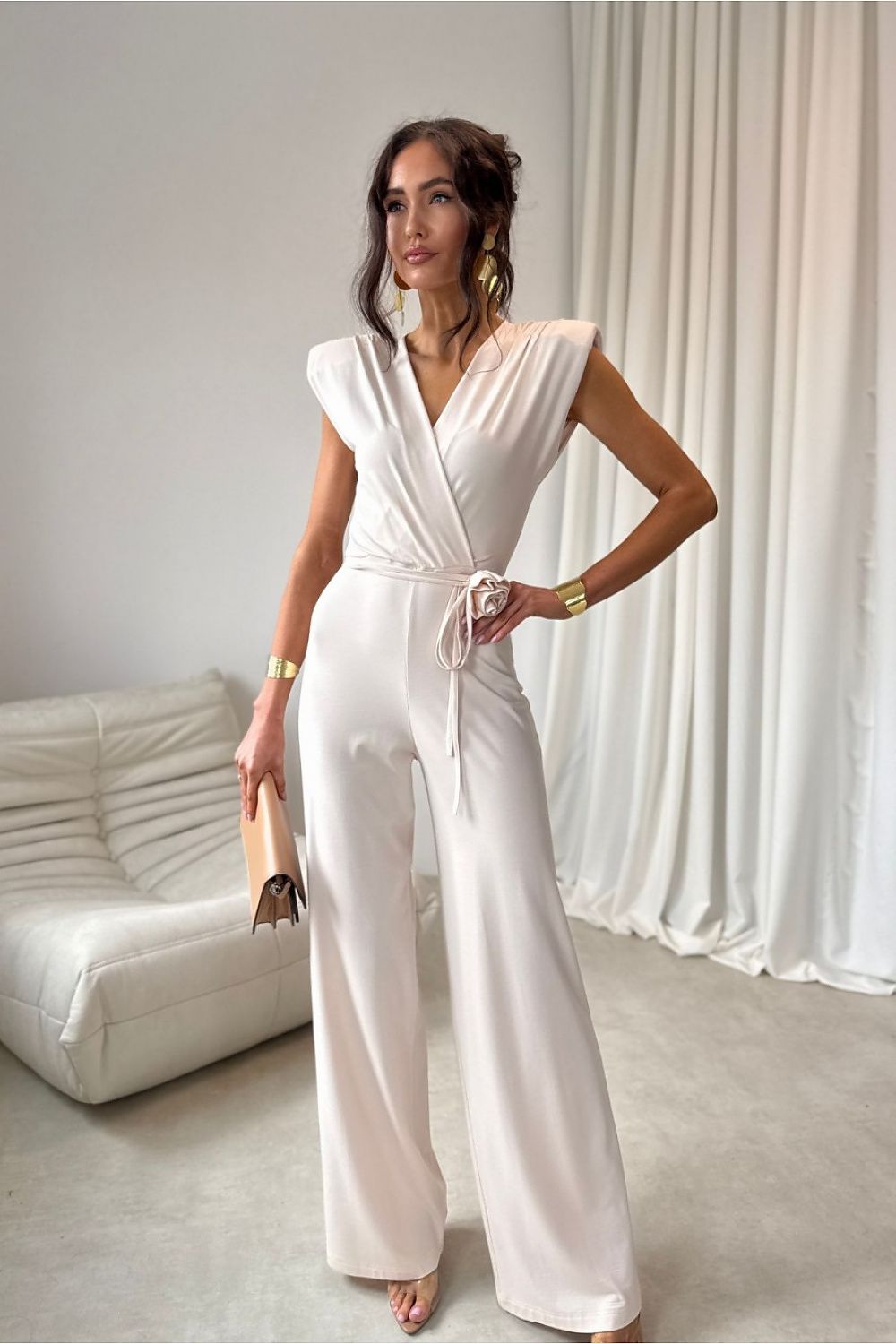 Woman in a white jumpsuit standing in a room with a white couch and curtains.