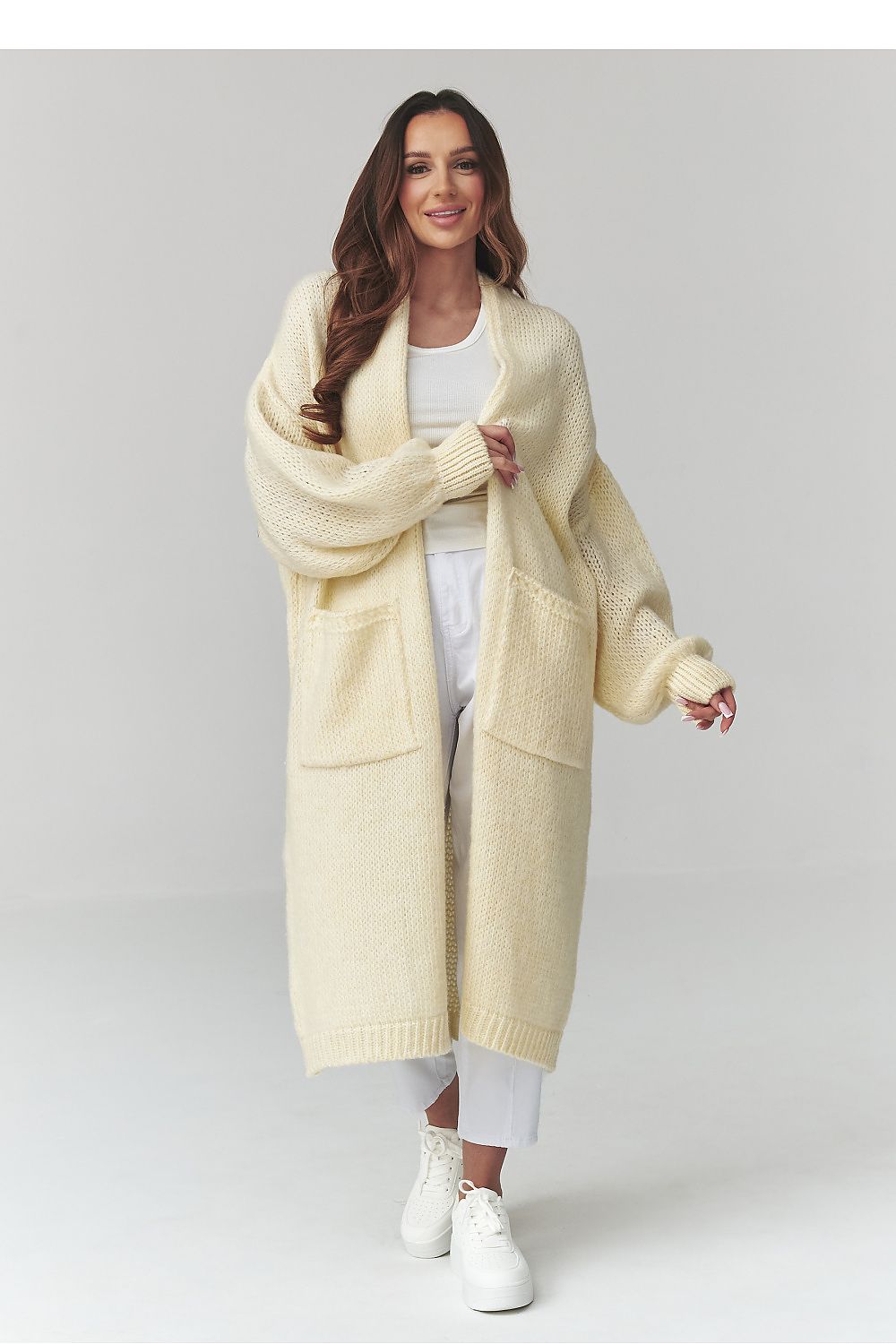 Woman wearing a long cream cardigan over a white outfit on a light gray background
