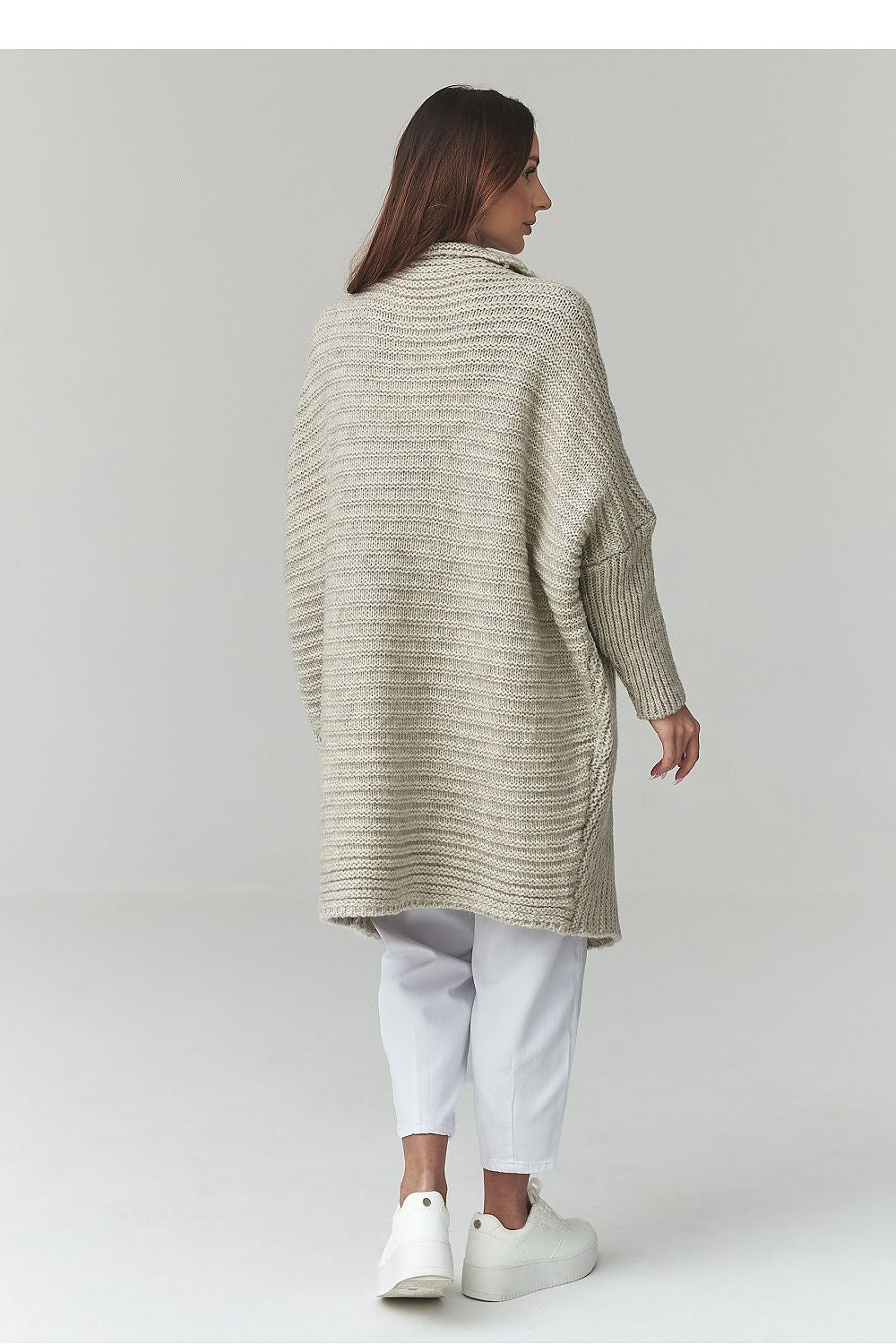 Woman wearing a long, light-colored cardigan over white pants on a plain background