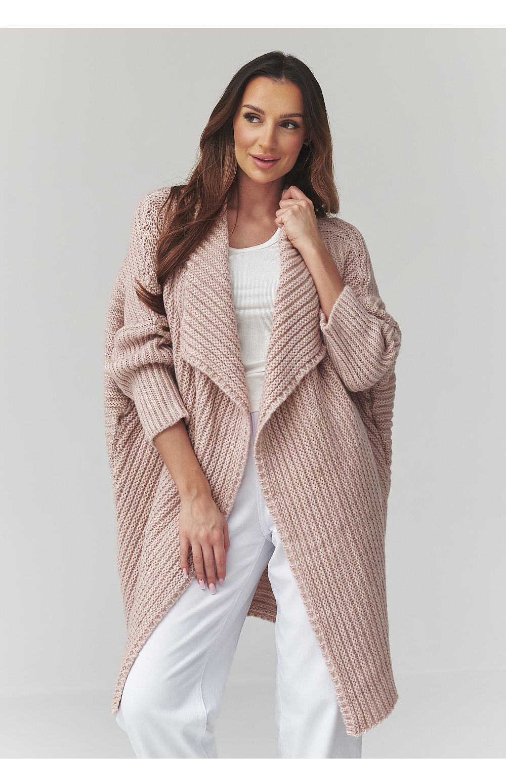 Woman wearing a long, light pink knitted cardigan over a white top and pants on a plain background