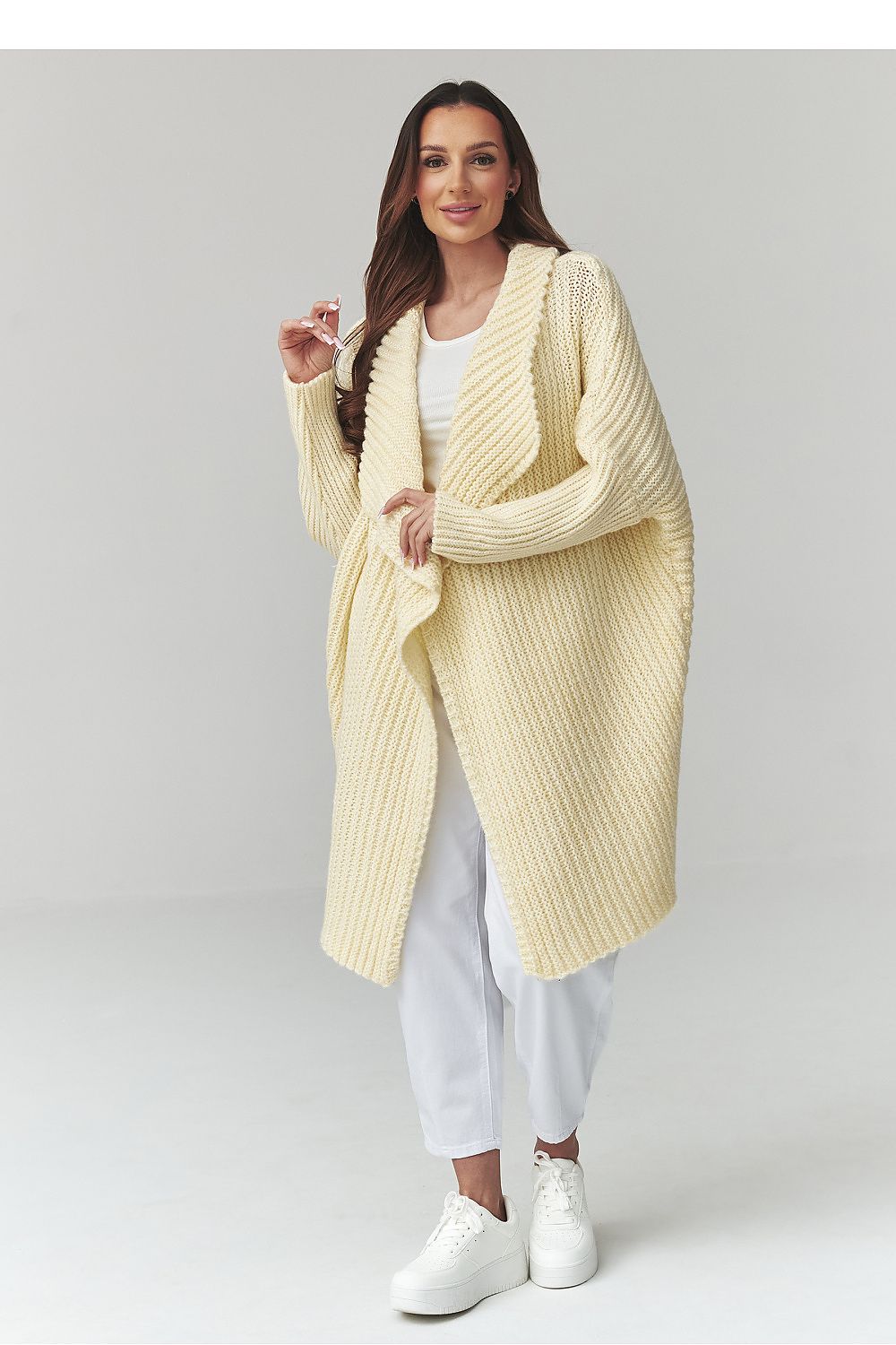 Woman wearing a long cream cardigan over a white outfit on a light gray background