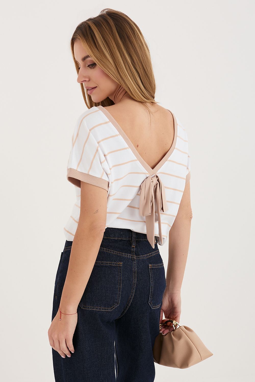 Woman wearing a white striped shirt with an open back and dark jeans, holding a beige clutch.