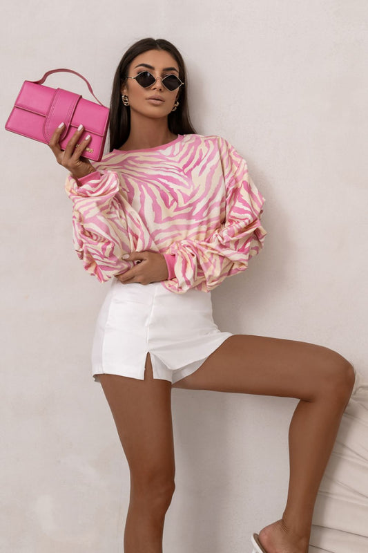 Woman wearing a pink and white patterned top with white shorts, holding a pink clutch against a plain background.