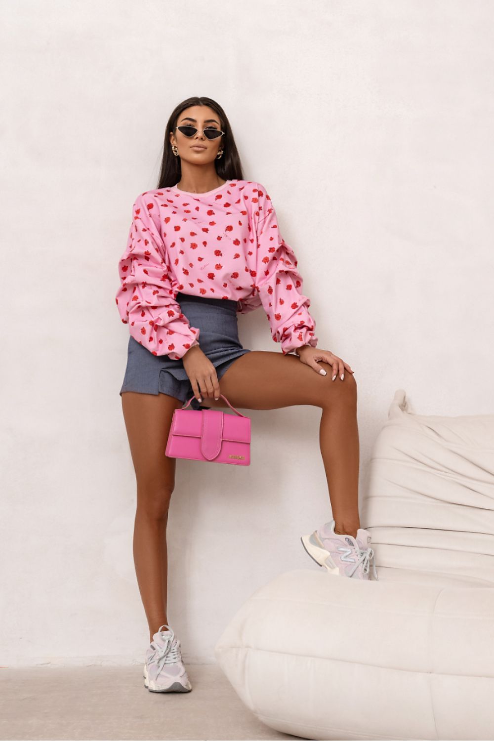 Woman wearing a pink heart-patterned sweatshirt and denim shorts, holding a pink handbag against a white background.