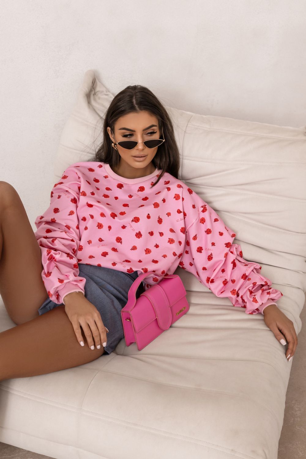 Woman wearing a pink sweatshirt with red patterns, sunglasses, and holding a pink handbag on a white couch.