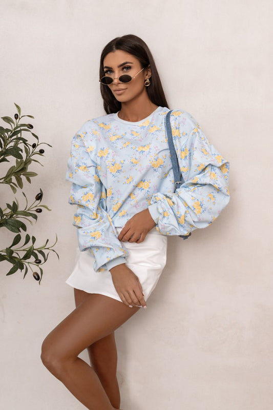 Woman wearing a light blue floral sweatshirt and white shorts against a beige wall.