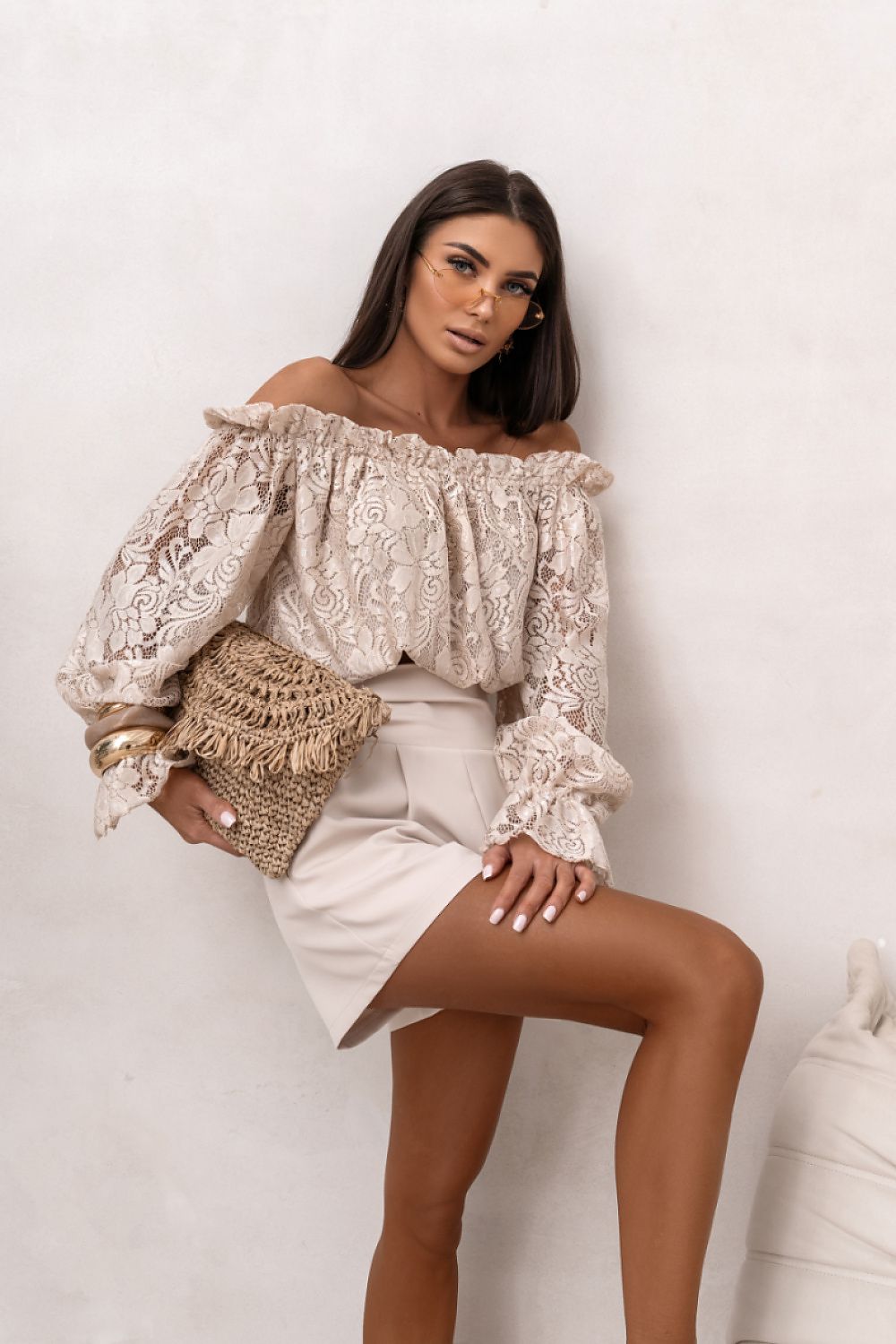 Woman wearing a beige off-shoulder top and white skirt, holding a woven clutch against a white background.
