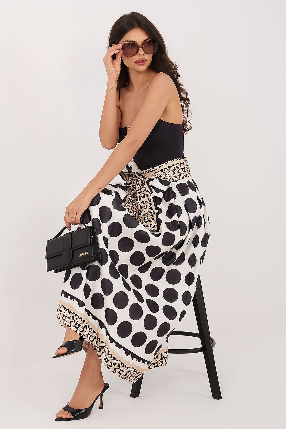 Woman wearing a black and white polka dot skirt with leopard print trim, holding sunglasses and a black handbag.