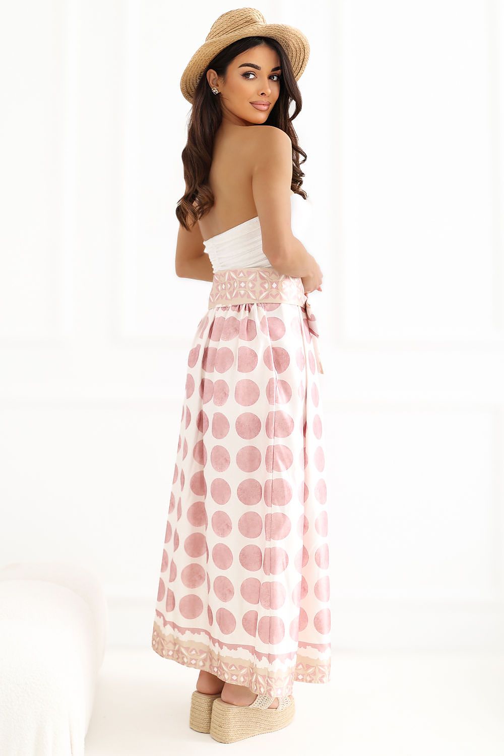Woman wearing a strapless white top and pink polka dot skirt with a straw hat on a white background