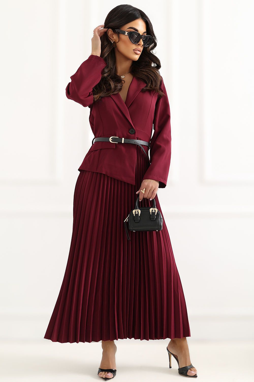 Woman wearing a burgundy suit with a pleated skirt and holding a black handbag on a white background
