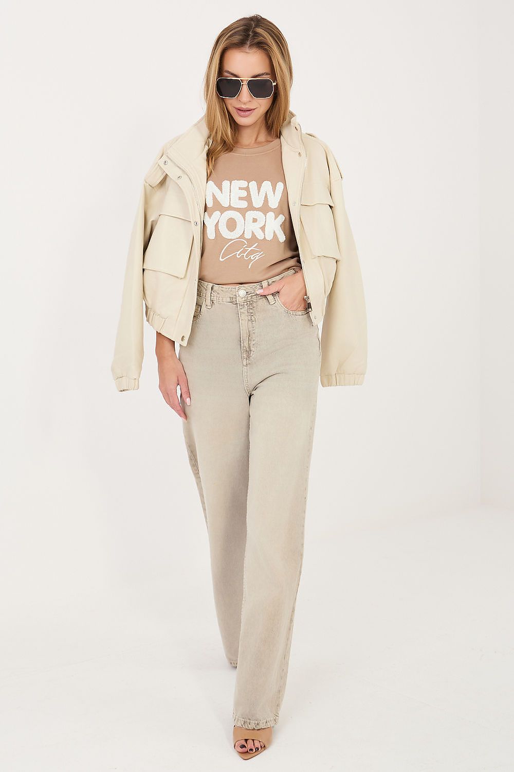 Woman wearing a beige jacket, graphic t-shirt, and jeans on a white background