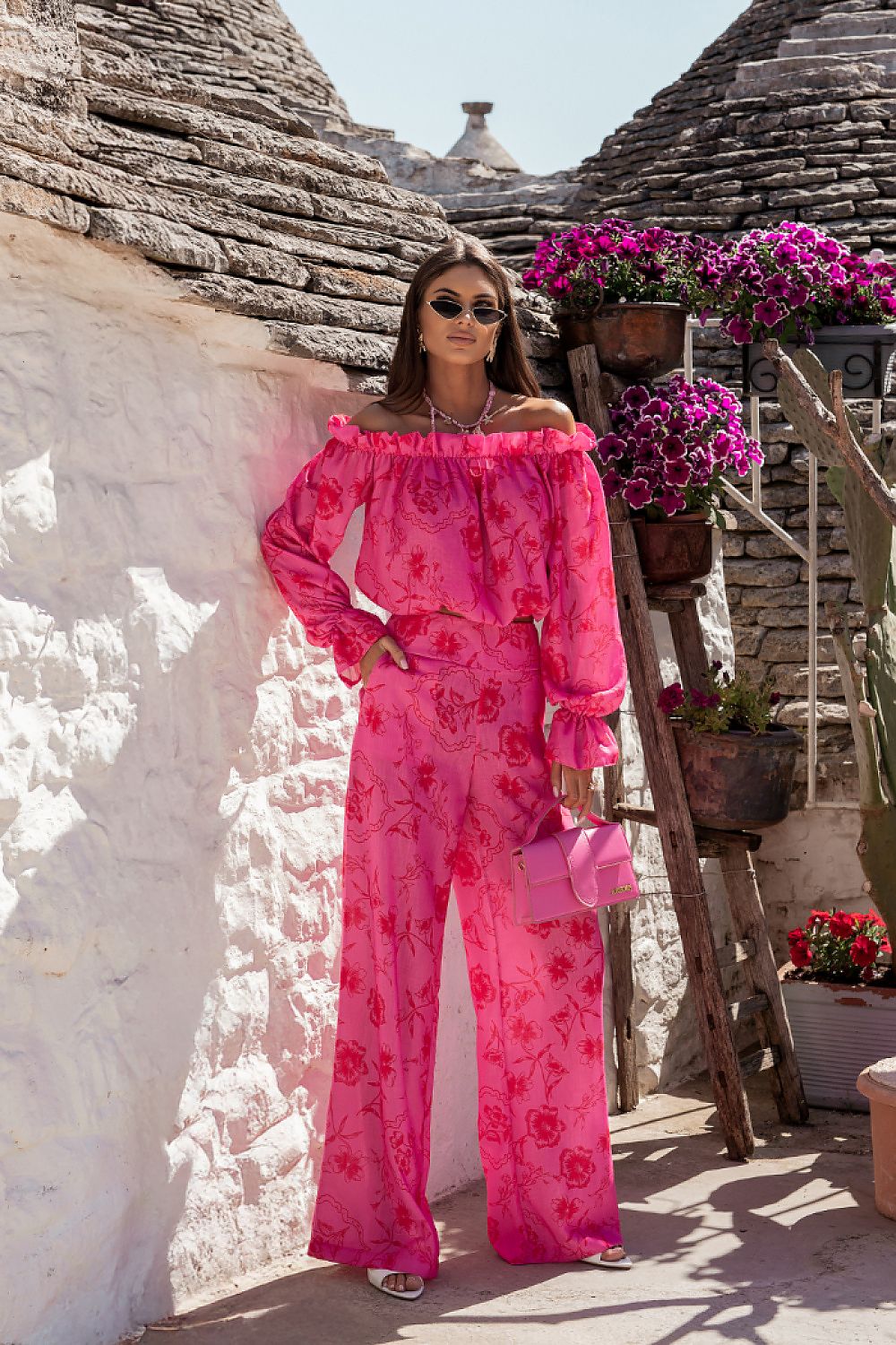 Woman in a bright pink floral outfit standing in front of stone huts with flowers.