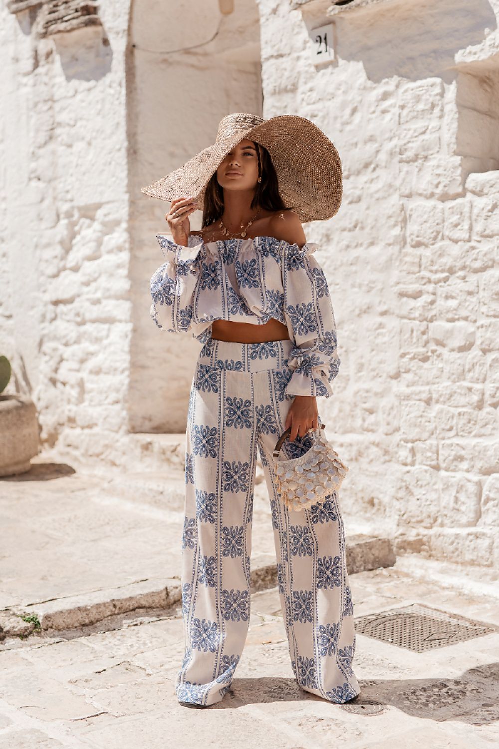 Woman in a blue and white patterned outfit with a wide-brimmed hat standing in front of a stone wall.