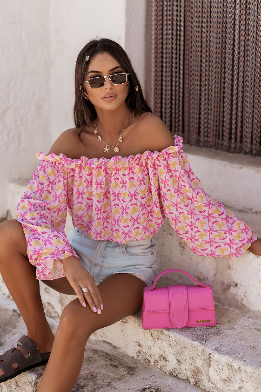 Woman in a floral off-shoulder top and denim shorts with a pink handbag, sitting on stone steps.