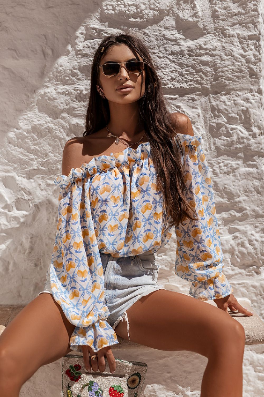Woman in a floral off-shoulder top and shorts sitting against a stone wall.