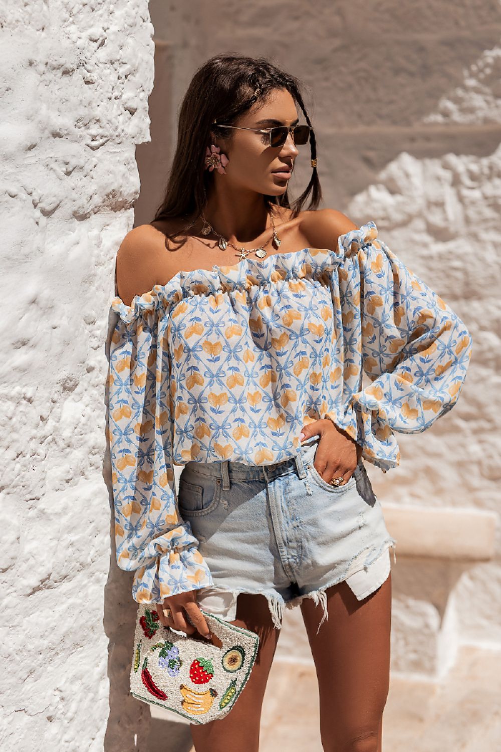 Woman wearing a floral off-shoulder top and denim shorts, holding a colorful handbag against a stone wall.