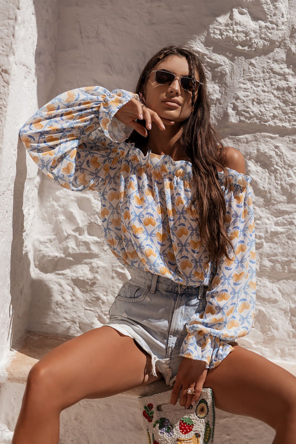 Woman in a floral blouse and denim shorts sitting against a textured wall.