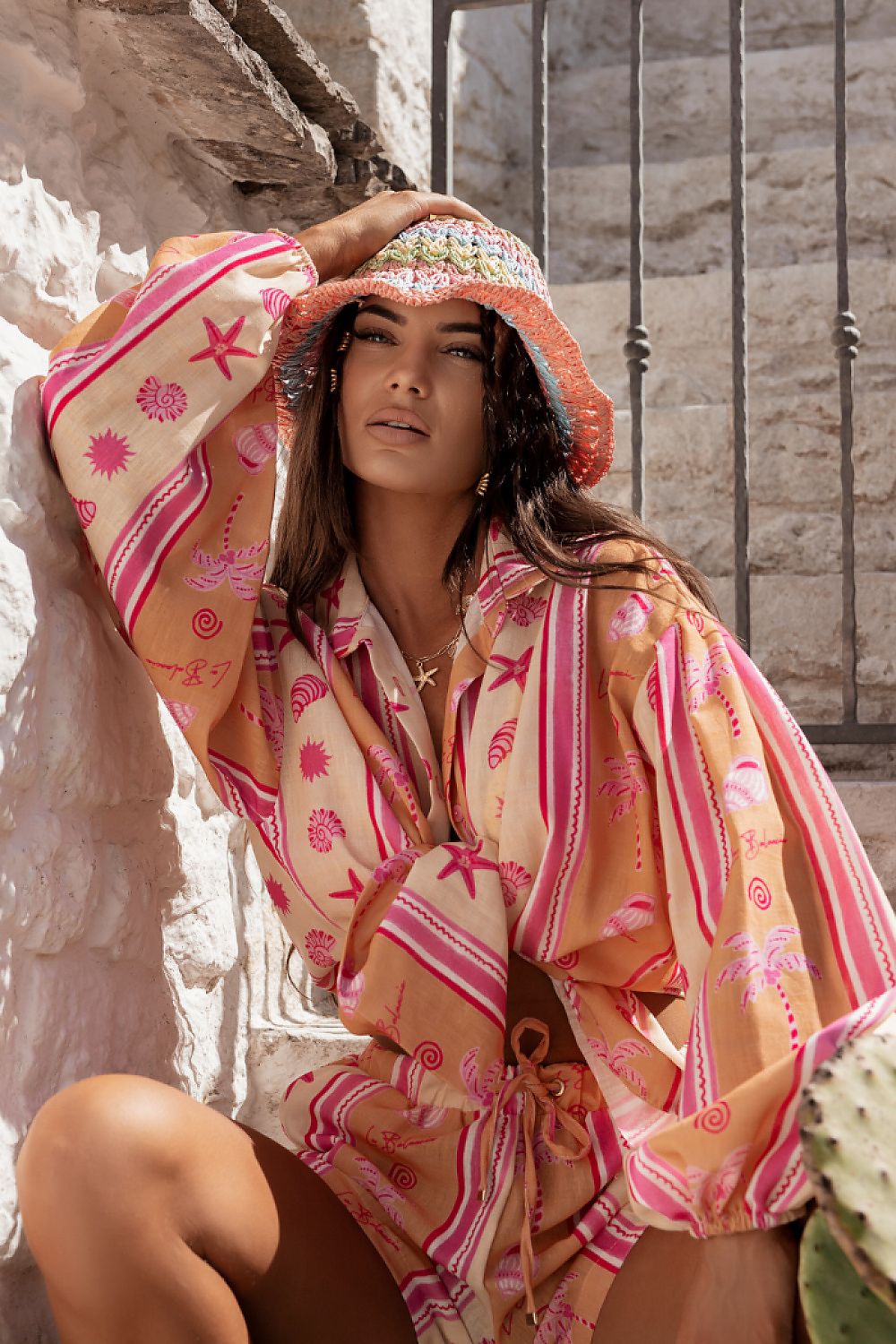 Woman wearing a patterned dress and hat against a stone wall.