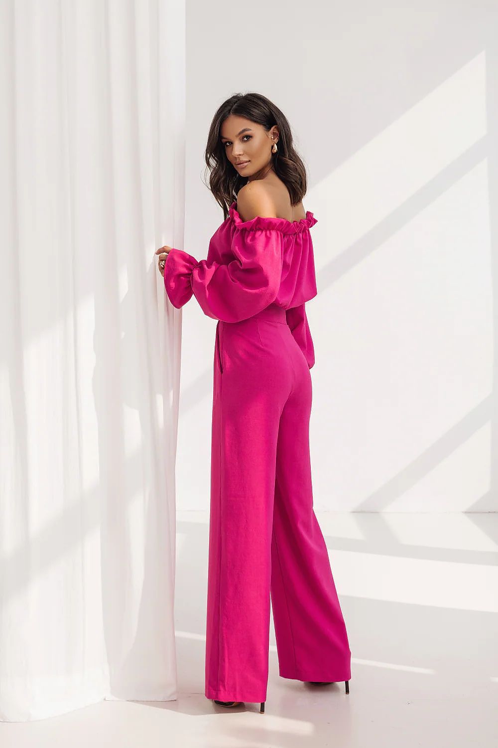 Woman wearing a bright pink off-shoulder top and wide-leg pants against a white curtain background.