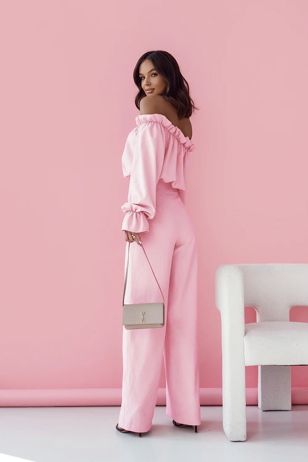 Woman in a pink off-shoulder outfit holding a handbag against a pink wall.