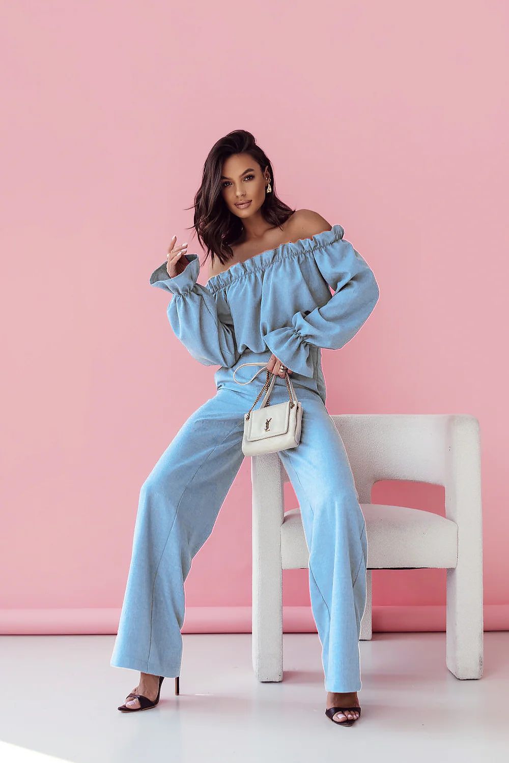 Woman in light blue off-shoulder top and pants sitting on a white chair against a pink background